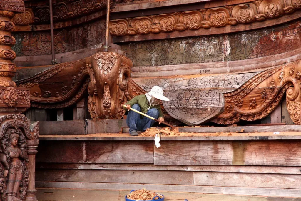 Restorer wearing a white hat carefully cleans intricate wooden carvings on an ancient temple.

