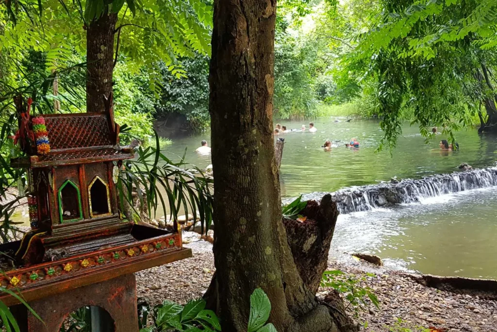 Small traditional shrine by a tree near a river with people swimming and a small waterfall