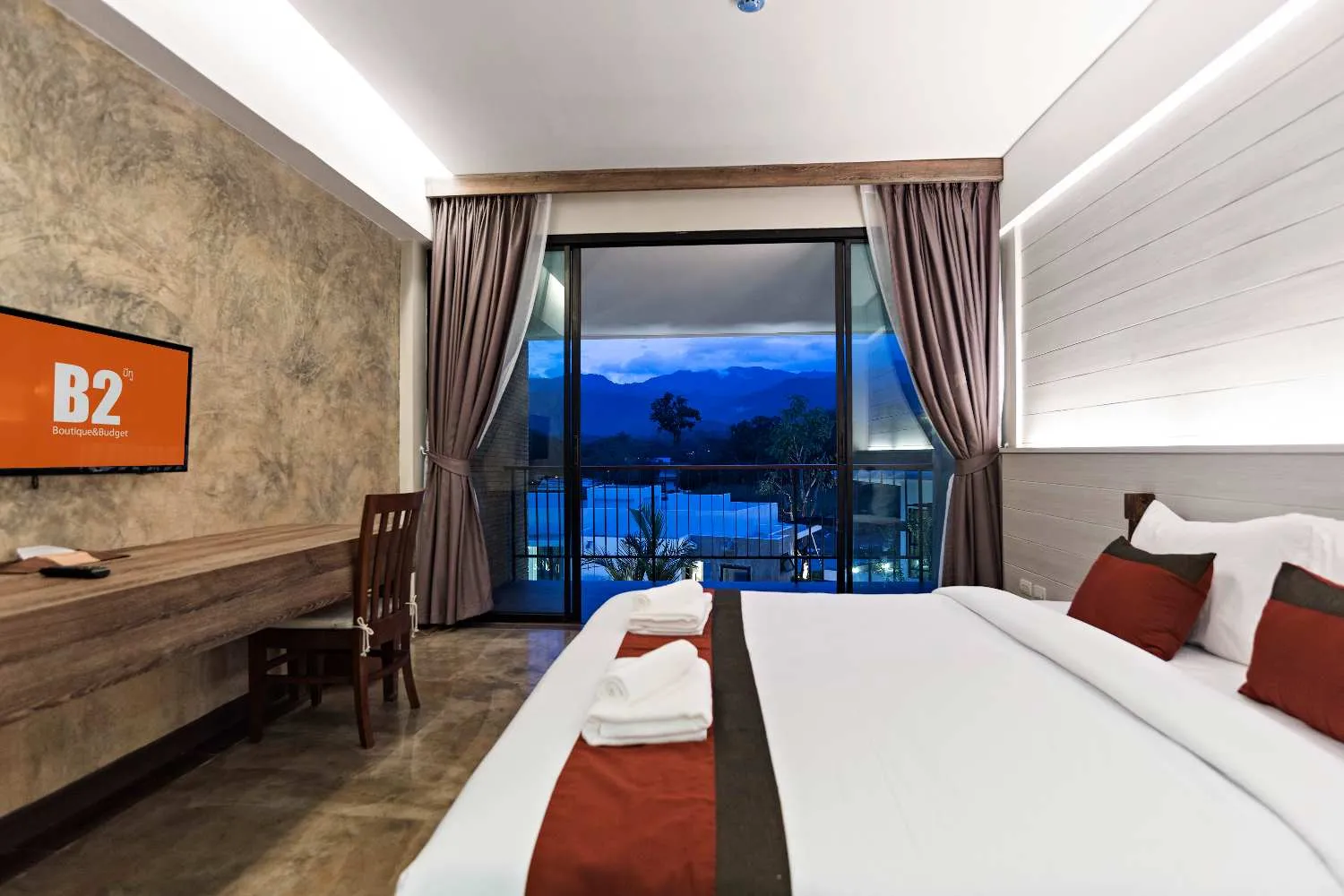 Modern hotel room with large bed, desk, and balcony overlooking a blue mountain landscape at dusk