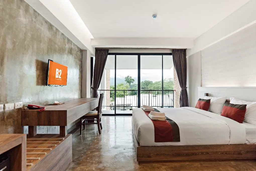 Modern hotel room with large bed, wooden furniture, and balcony overlooking greenery and water