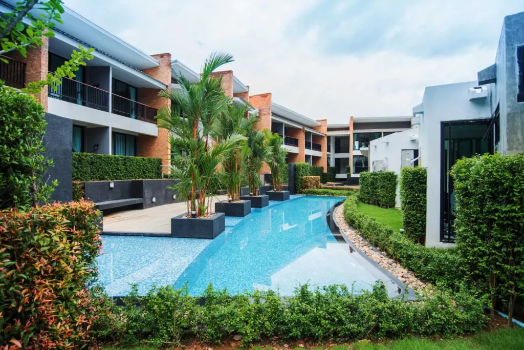 Modern resort with a long swimming pool, palm trees, and contemporary balconies under a cloudy sky