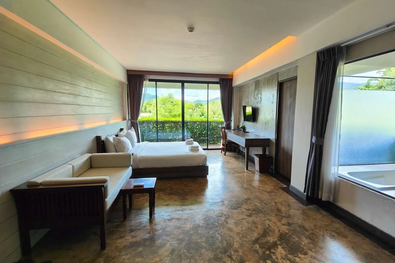 Bright hotel room with large bed, seating area, desk, and window view of green trees.