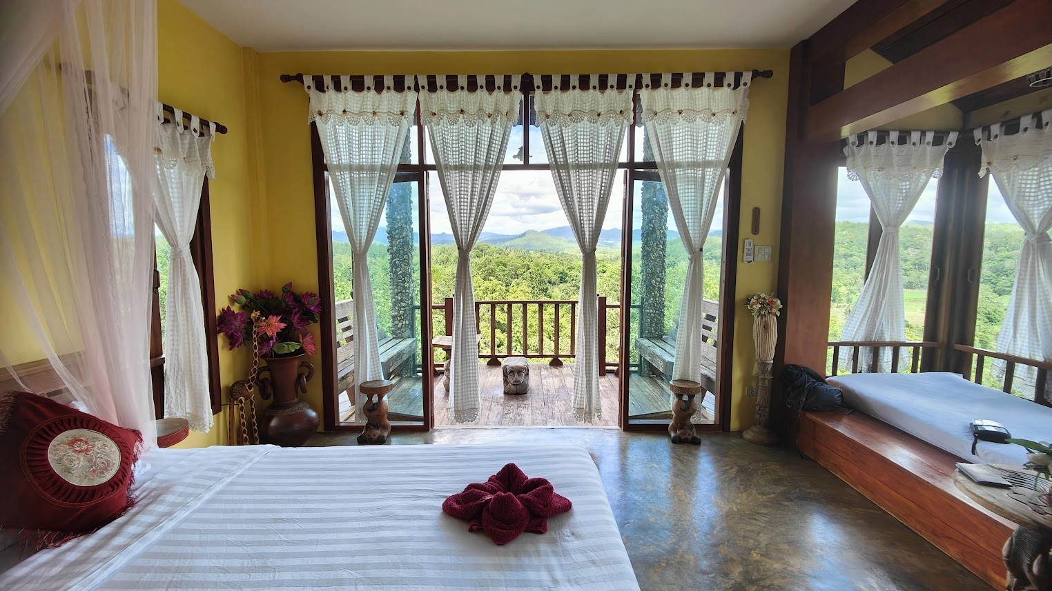 Sunlit bedroom with open glass doors leading to a balcony overlooking green hills and trees.

