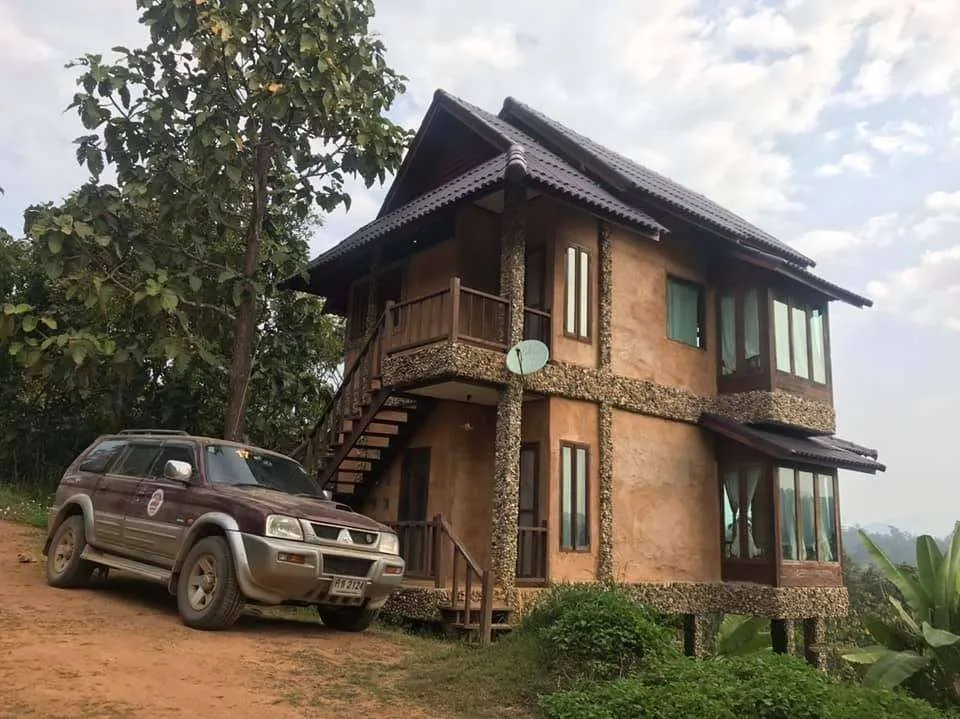 Two-story rustic brick house on a hillside with a brown SUV parked beside it under a cloudy sky

