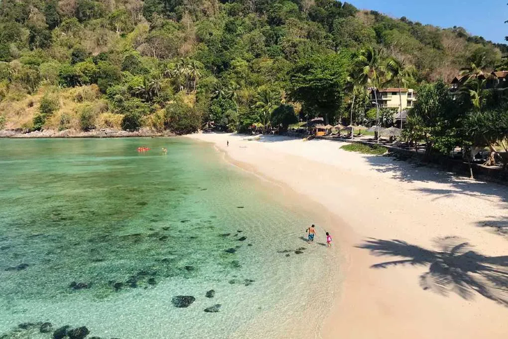 Clear turquoise ocean water meets white sandy beach with two people walking near shore and forested hills in background

