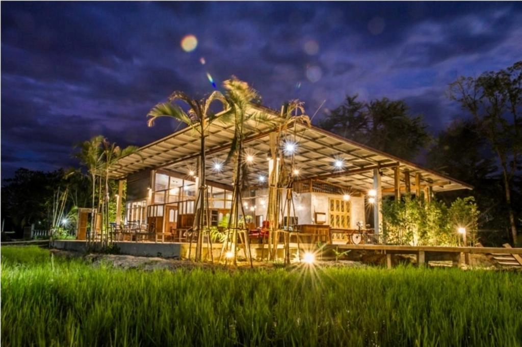 Modern wooden tropical resort lit up at dusk with palm trees and lush green grass surrounding it

