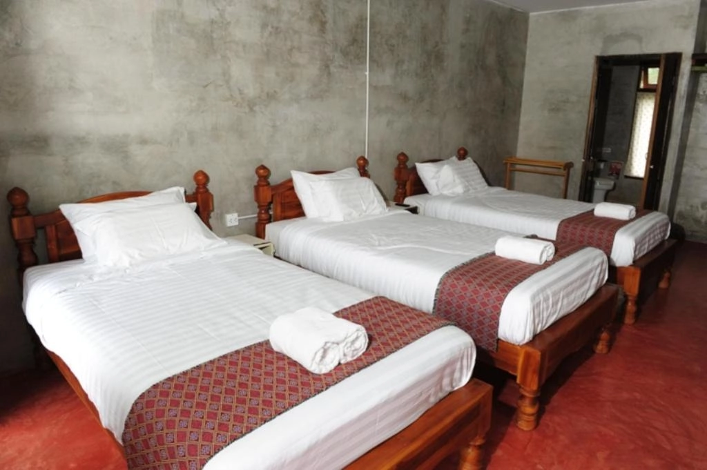 Three single beds with white linens and folded towels in a simple concrete-walled room.

