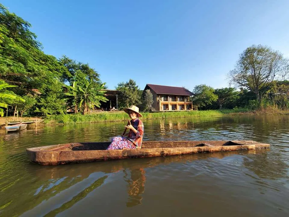 Woman wearing a hat sitting on a wooden boat floating on a calm river near green trees and houses.