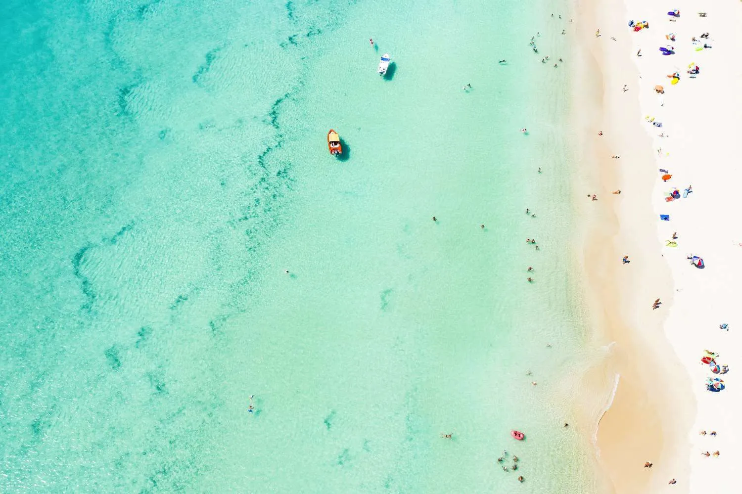 Aerial view of turquoise ocean water and white sandy beach with scattered umbrellas and boats
