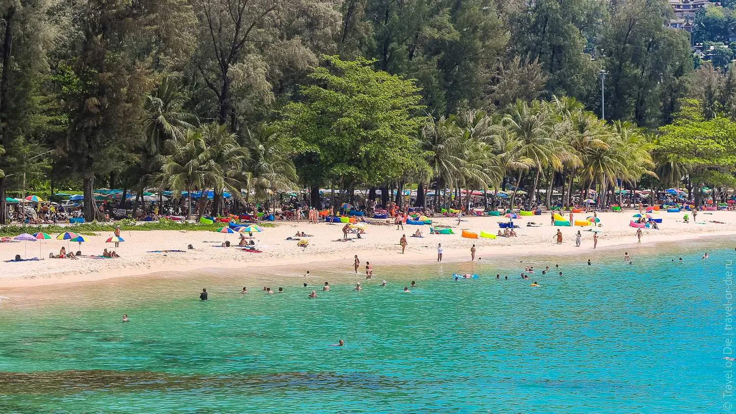 Crowded tropical beach with turquoise water, sunbathers, and lush green trees in background