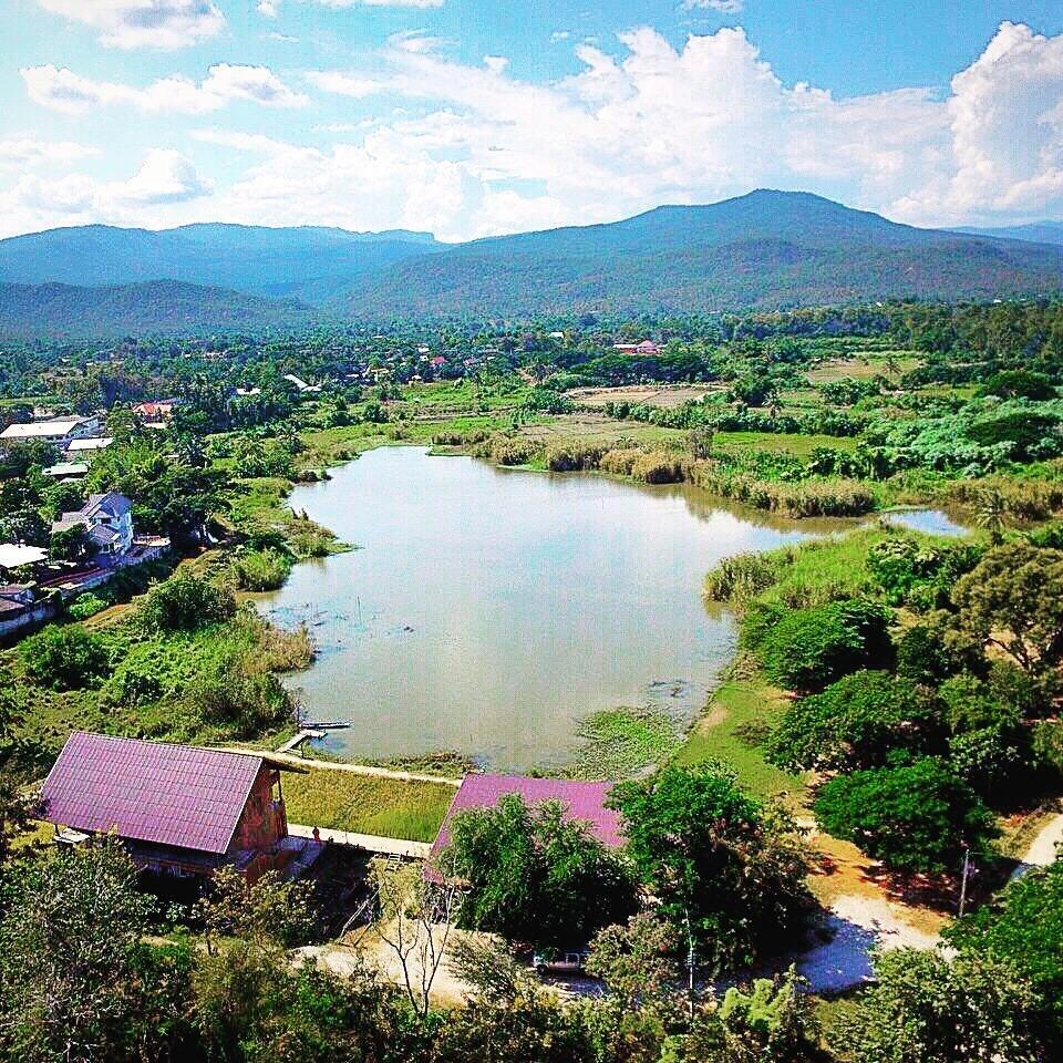 Aerial view of a serene lake surrounded by green fields and distant mountains under a partly cloudy sky.

