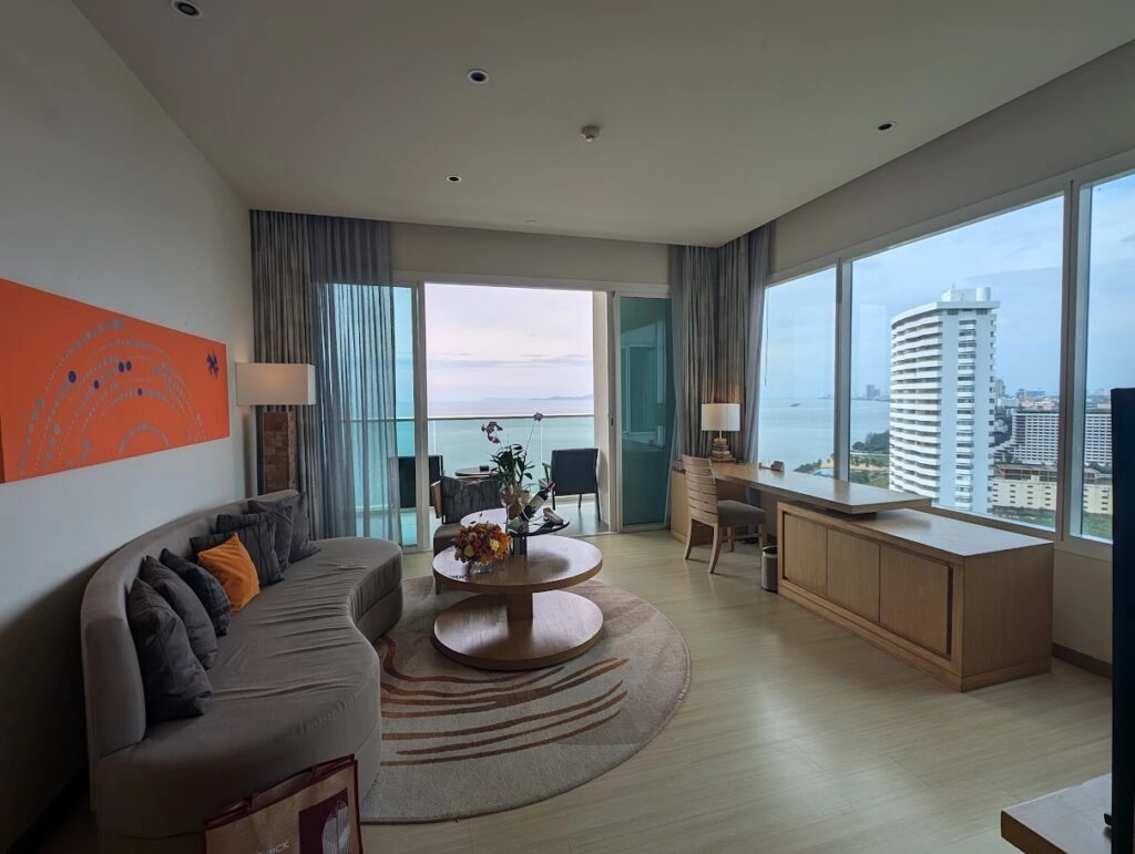 Spacious hotel room with modern furniture and ocean view from large windows in a cityscape.