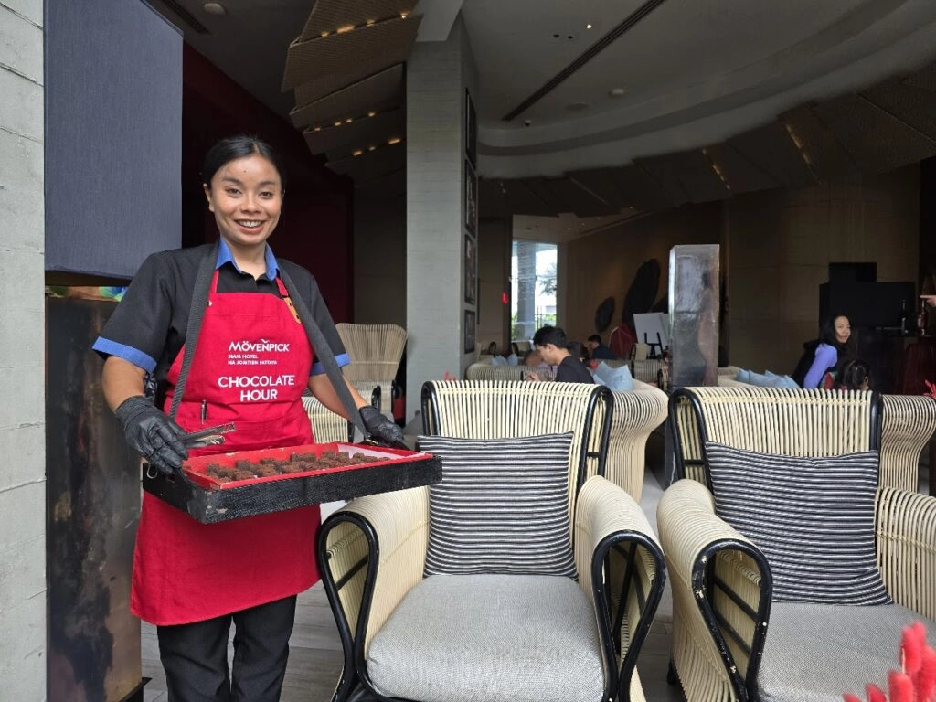 Smiling woman in red apron holding chocolate tray in a cozy cafe with armchairs