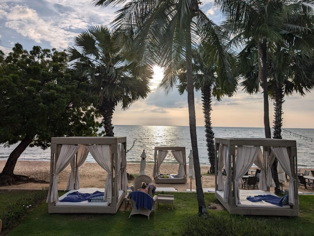 Beachside cabanas with white curtains and blue cushions under palm trees at sunset.
