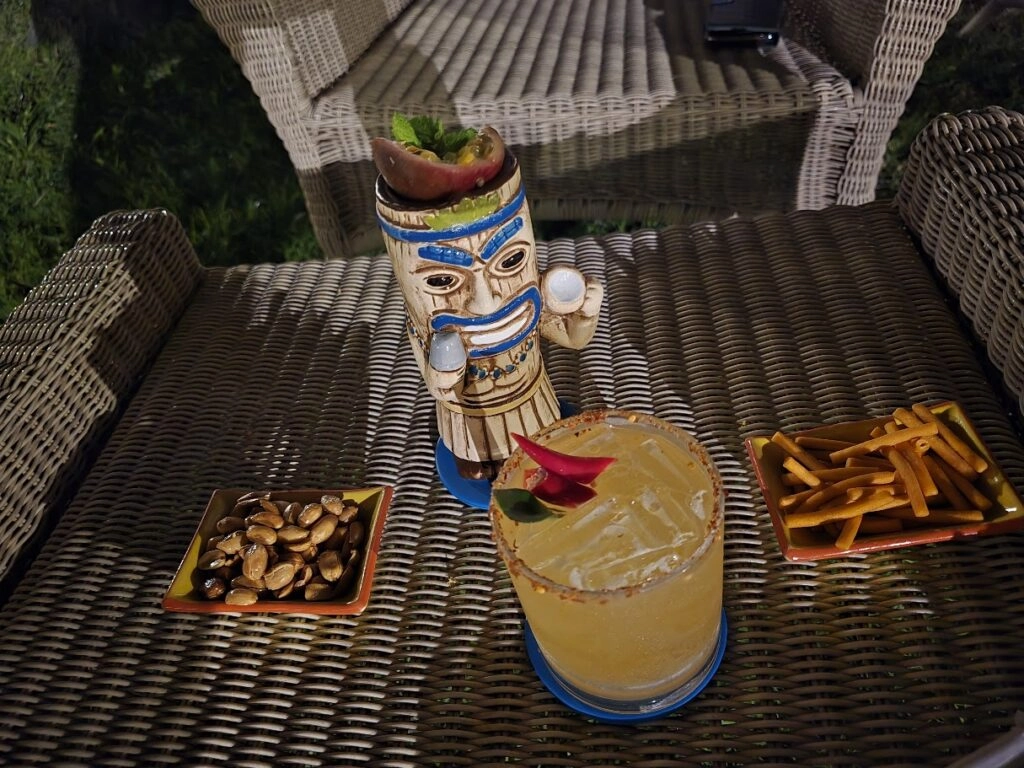 Tiki mug with a drink, snacks, and wicker chairs on an outdoor table at sunset