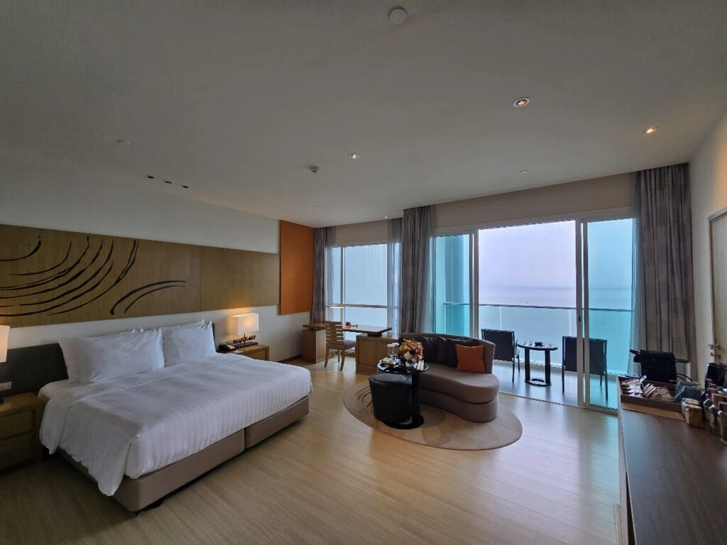 Modern hotel room with king bed, seating area, and large windows overlooking the ocean.