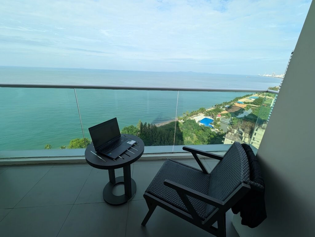 Laptop on a small table beside a chair on a balcony overlooking a calm ocean and coastline.