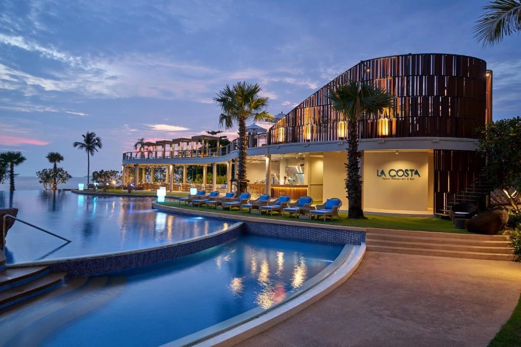 Modern beachfront hotel with palm trees and infinity pool at sunset, clear sky.