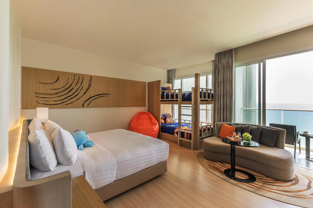 Modern hotel room with double bed, bunk beds, seating area, and ocean view balcony.