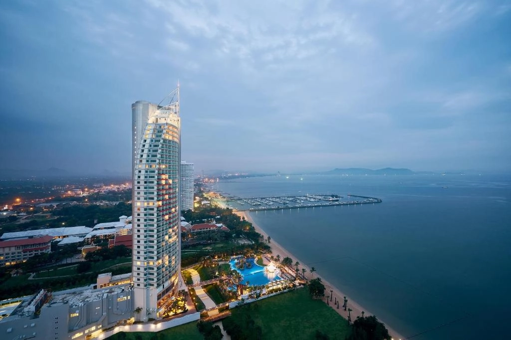Tall modern hotel building next to beach with pools and city lights at dusk