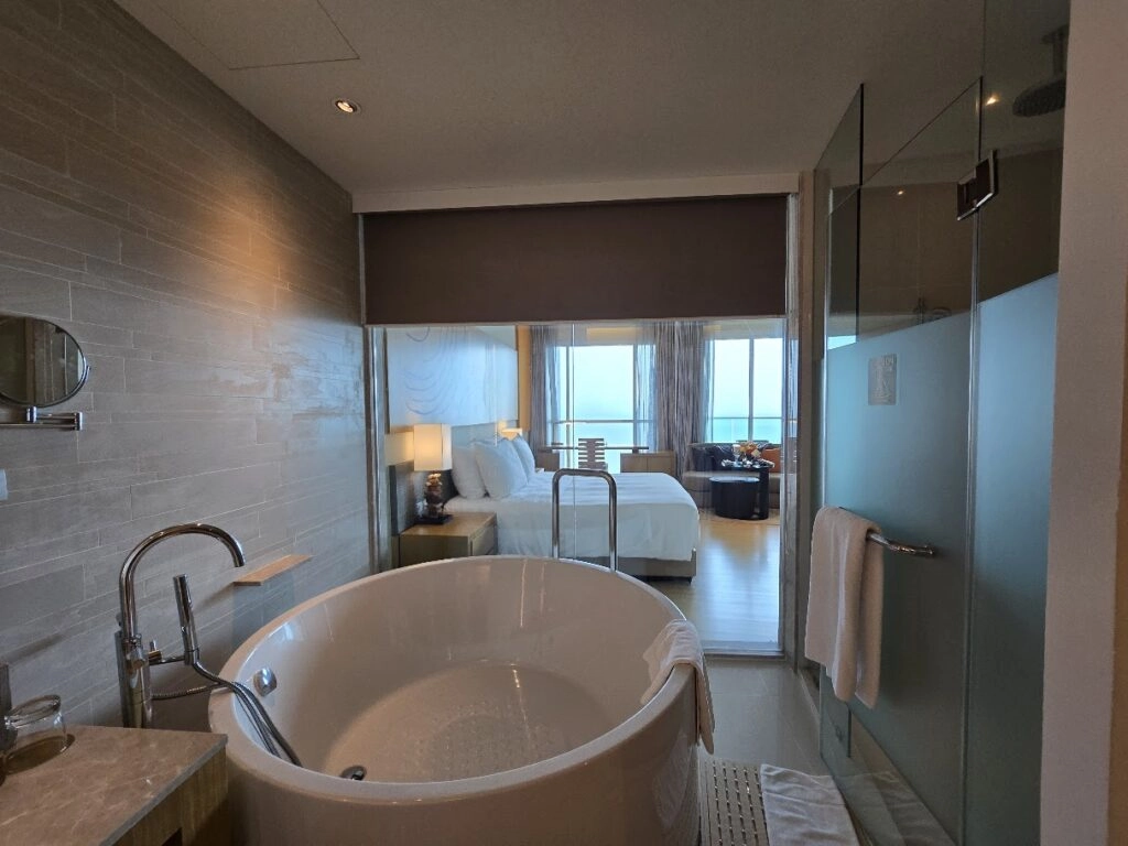 Modern hotel bathroom with round bathtub, glass shower, and bedroom with ocean view in background