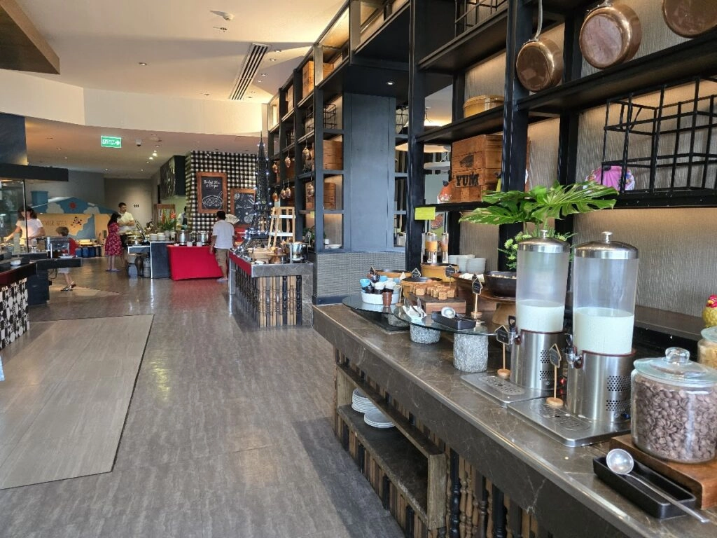 Modern hotel breakfast buffet area with cereal dispensers, pastries, and seating in background