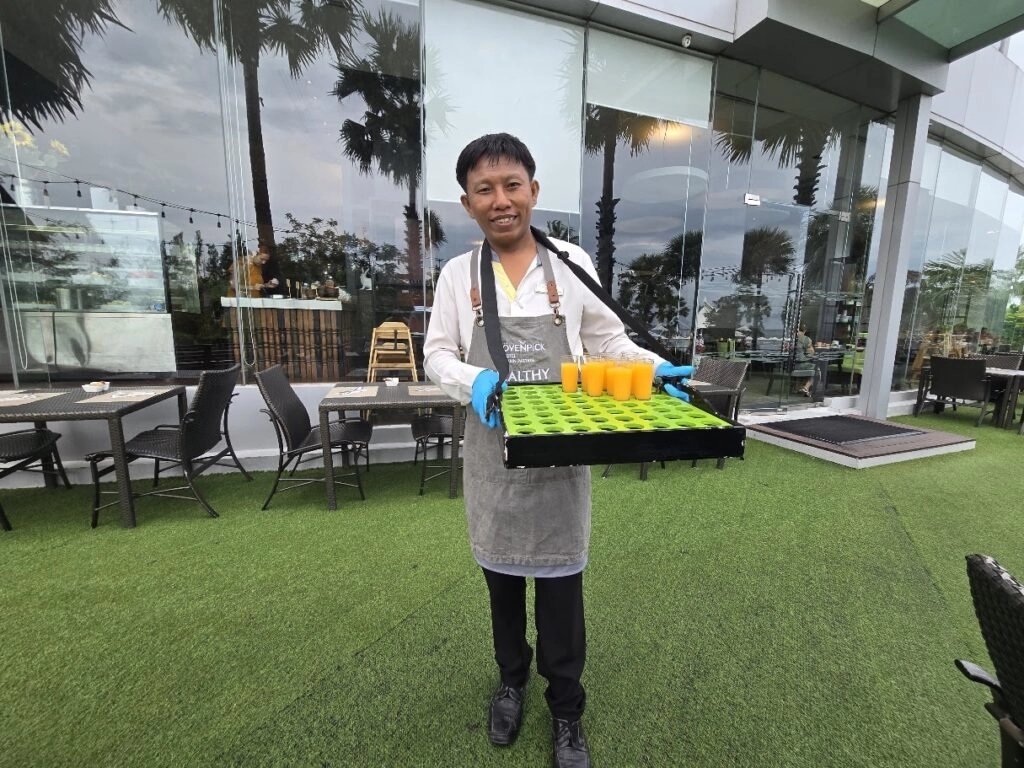 Young man holding tray of orange and green drinks outside modern cafe with glass walls