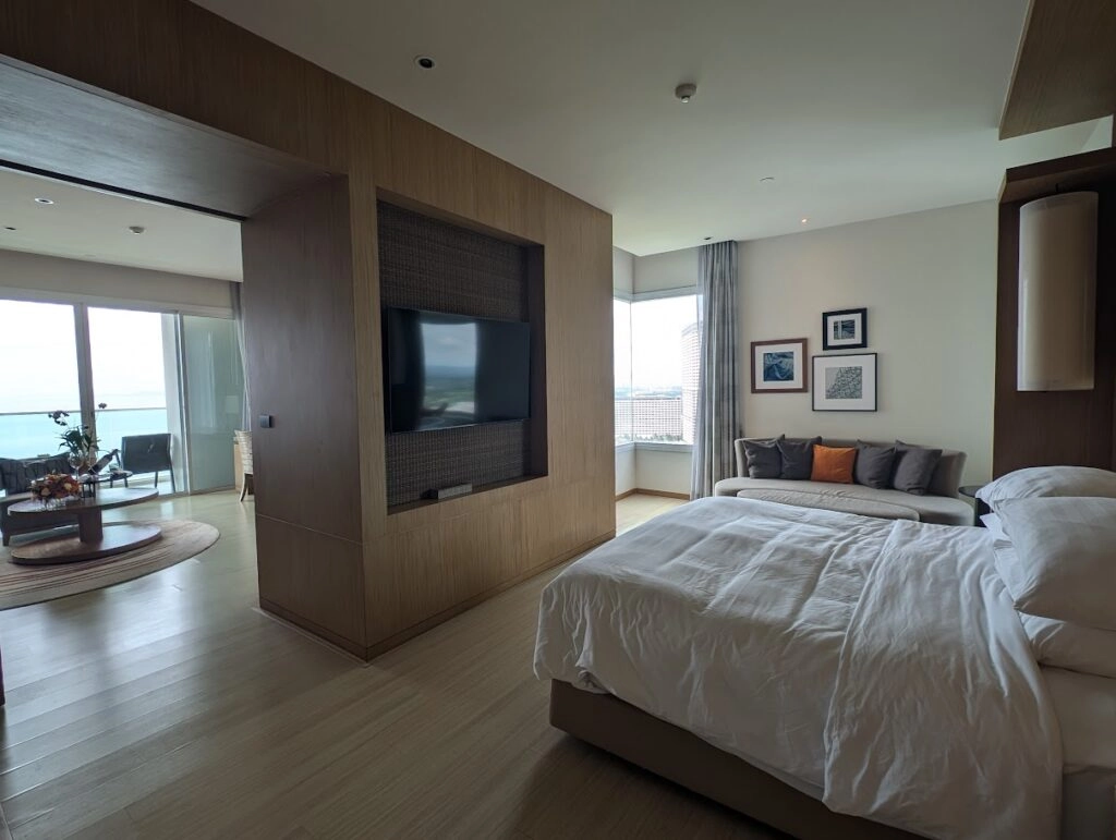 Modern hotel room with large bed, TV, seating area, and ocean view through balcony doors