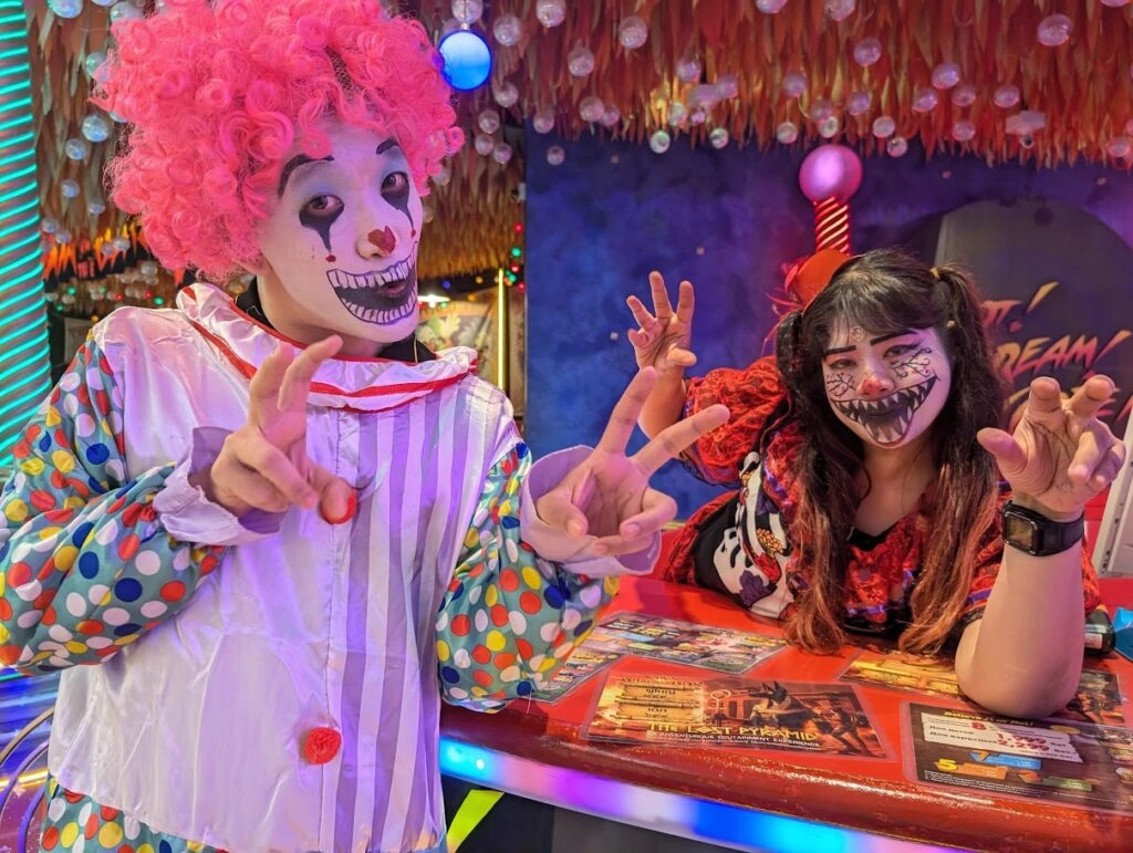 Clown with pink wig and colorful outfit posing with woman in spooky makeup at themed bar.