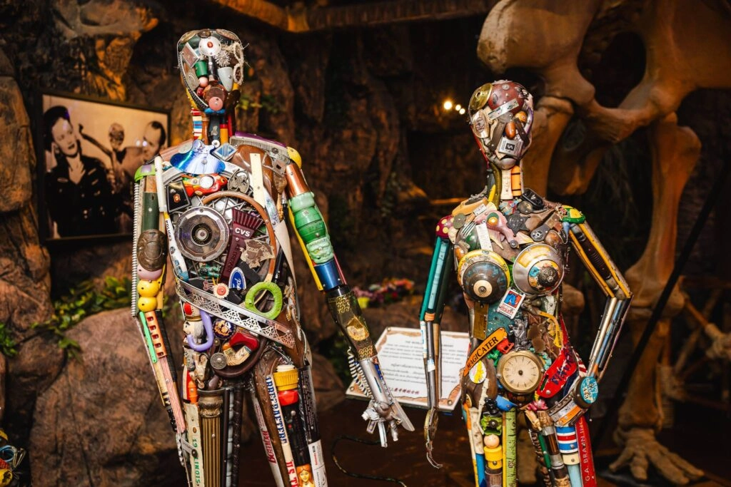 Two human-like sculptures made from colorful recycled electronic parts in a dim museum setting.