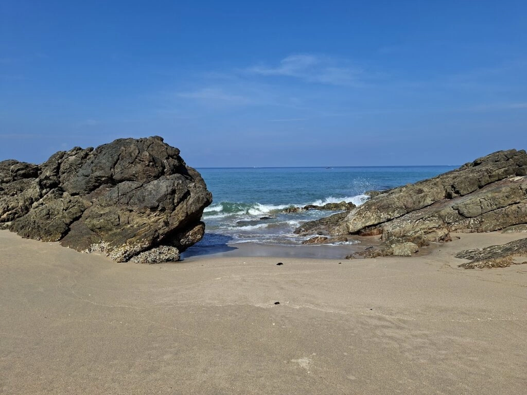 Zonovergoten zandstrand met golven tussen twee rotsformaties en blauwe hemel.