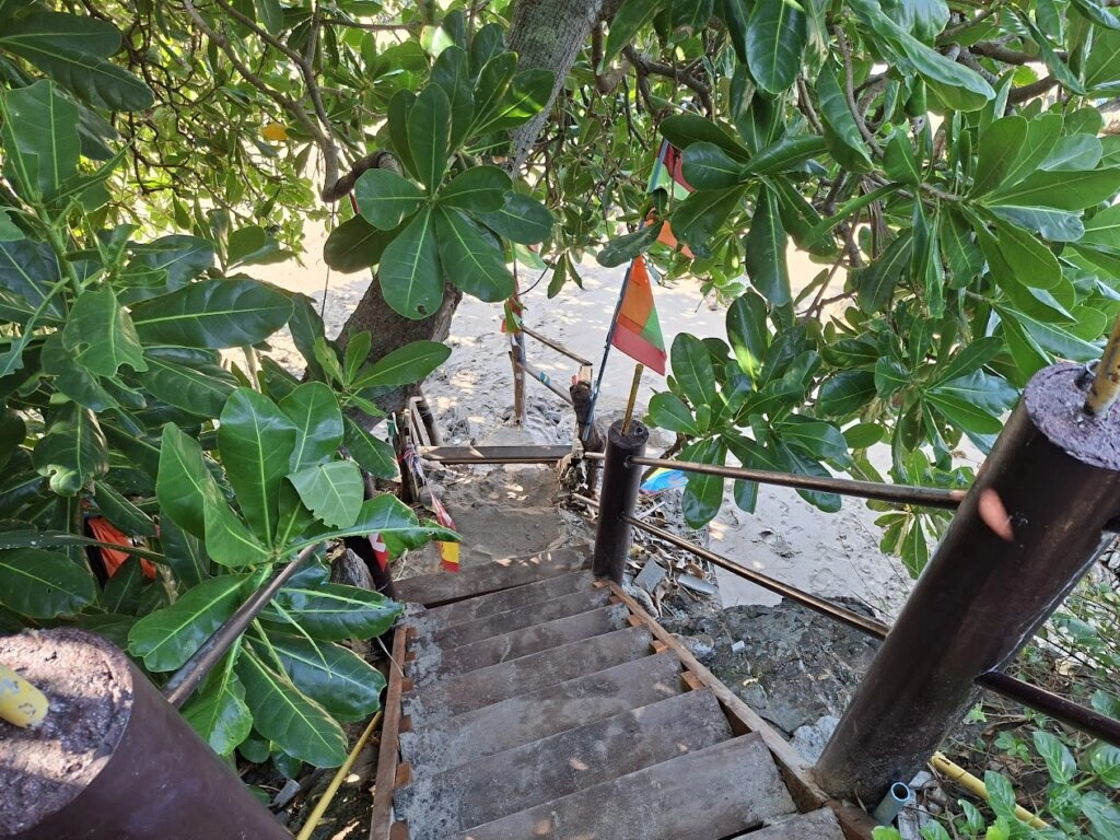 Houten trap naar een zandstrand tussen groene bladeren, met een rood-witte vlag.